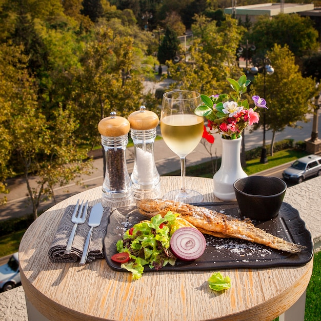 Seitenansicht gebratener Fisch mit Salat, Tomaten, Zwiebeln und Soße auf einem schwarzen Teller, ein Glas Weißwein auf einem kleinen runden Tisch mit Blick auf die Stadt