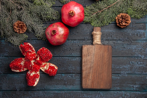 Kostenloses Foto seitenansicht granatäpfel auf tisch pilled granatapfel neben zwei roten granatäpfeln holzbrett und fichtenzweigen mit zapfen