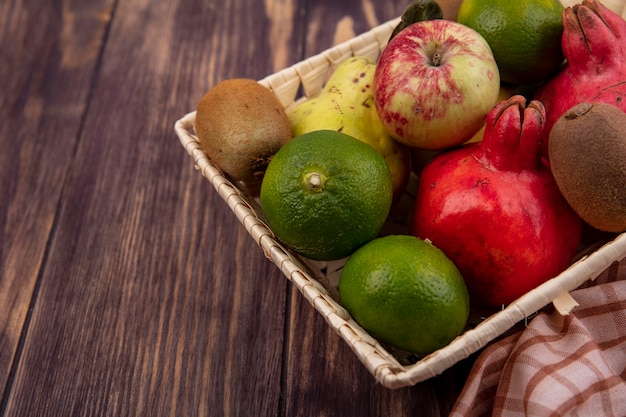 Seitenansicht Granatäpfel mit Mandarinenäpfeln und Kiwi in einem Korb auf einer Holzwand