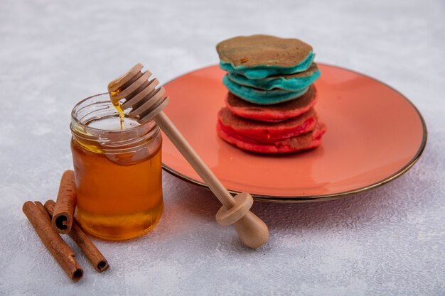 Seitenansicht Honig in einem Glas mit einem Holzlöffel Zimt und bunten Pfannkuchen auf einem orangefarbenen Teller auf einem weißen Hintergrund