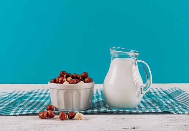 Seitenansicht-Milchkaraffe mit Schüssel Mandeln auf weißem hölzernem und blauem Stoffhintergrund. horizontaler Raum für Text