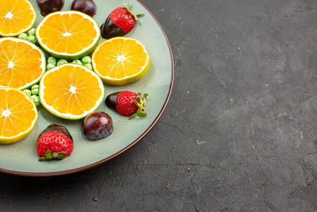 Seitenansicht mit Schokolade überzogene erdbeergrüne Bonbons gehackte orange und mit Schokolade überzogene Erdbeeren auf dem Teller auf der linken Seite des schwarzen Tisches