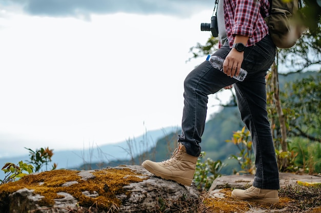 Seitenansicht Nahaufnahme der Beine eines jungen asiatischen Wanderers, der die Kamera auf dem Knie steht und eine Wassergetränkeflasche mit glücklich auf dem Gipfel des felsigen Berges hält schöne Landschaft im Hintergrund Kopierraum