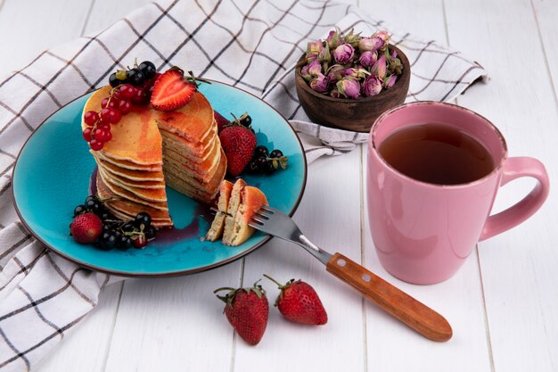 Seitenansicht Pfannkuchen mit Erdbeeren der schwarzen und roten Johannisbeeren mit einer Gabel auf einem Teller mit einer Tasse Tee auf einem weißen karierten Handtuch