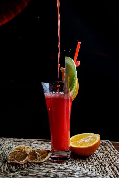 Seitenansicht roter Fruchtsmoothie mit Tubuli für Getränke und halbe Orange und Tropfen in Servietten