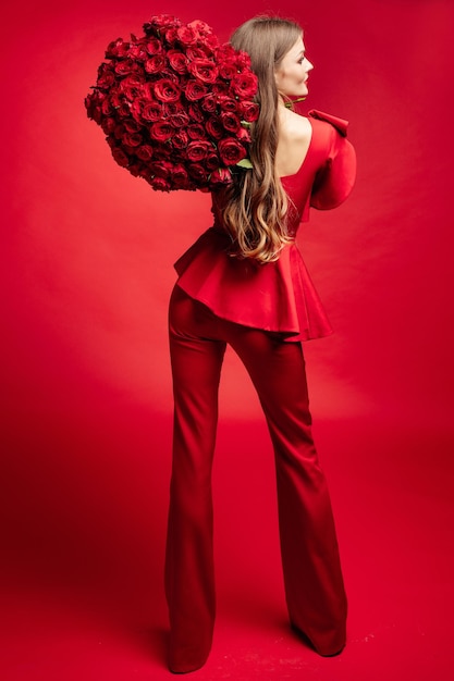 Seitenansicht Studioporträt einer wunderschönen jungen Brünette mit langen Haaren, die ein modisches rotes Kleid trägt. Sie lächelt einen großen Strauß roter Rosen in ihren Händen an. Total rote Farbe auf rotem Hintergrund isolieren