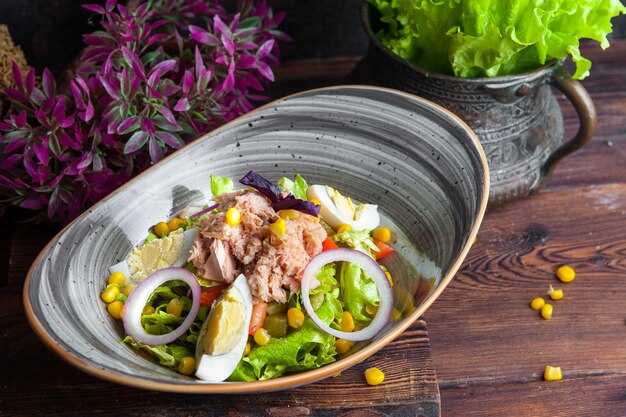 Seitenansicht Thunfischsalat mit Salat, Eiern, Tomaten, Gurke, Zwiebel und Mais auf einem dunklen Holztisch horizontal