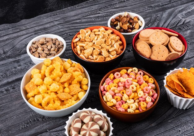 Seitenansicht verschiedener Arten von Snacks als Nüsse, Cracker und Kekse in Schalen auf dunkler Oberfläche horizontal