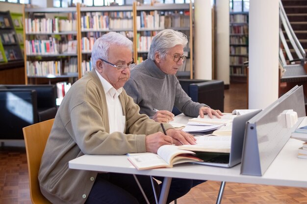 Seitenansicht von älteren Männern, die aktiv studieren. Zwei grauhaarige Männer mit Brille, die am Tisch saßen, konzentrierten sich darauf, mit Stiften Notizen zu machen und sich auf ihre Computerkurse vorzubereiten. Bildung für erwachsene Menschen Konzept