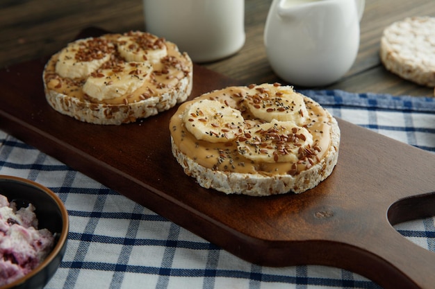 Kostenloses Foto seitenansicht von bananen-erdnussbutter-cripbread-snacks auf schneidebrett mit hüttenkäsemilch geronnener milch auf kariertem stoff auf holzhintergrund