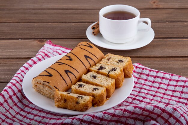 Seitenansicht von Biskuitkuchen mit Schokolade auf einem weißen Teller und einer Tasse Tee auf Holz