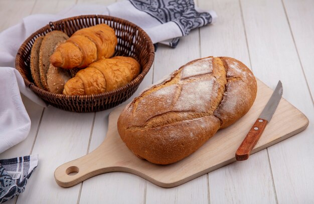 Seitenansicht von Broten als Croissant und gesäte braune Kolbenbrotscheiben im Korb auf Stoff und knusprigem Brot mit Messer auf Schneidebrett auf Holzhintergrund