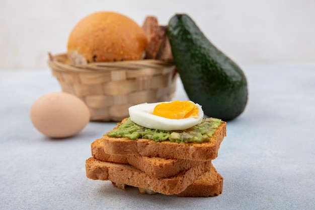 Seitenansicht von Broten mit Avocado-Fruchtfleisch und gekochtem Ei mit einem Eimer Brot mit Avocado und Ei auf weißer Oberfläche