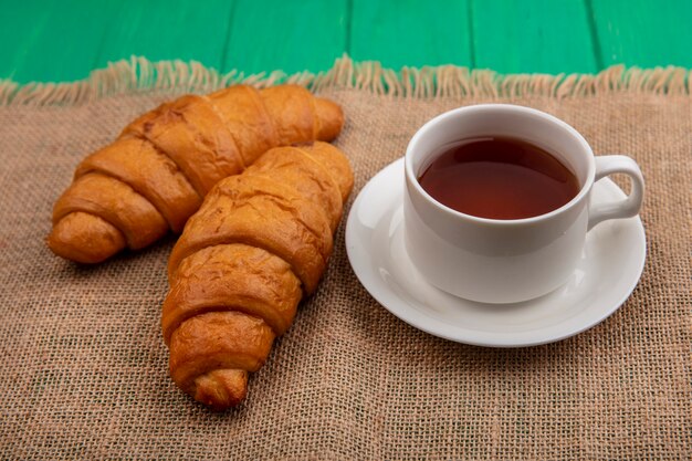 Seitenansicht von Croissants und Tasse Tee auf Untertasse auf Sackleinen auf grünem Hintergrund