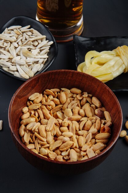 Seitenansicht von Erdnüssen in einer Holzschale und in Sonnenblumenkernen mit Streichkäse und einem Becher Bier auf Schwarz