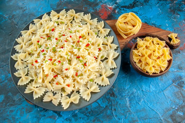 Seitenansicht von Farfalle-Nudeln mit Gemüse auf einem schwarzen Teller auf Holzbrett und verschiedenen Nudeln auf blauem Hintergrund