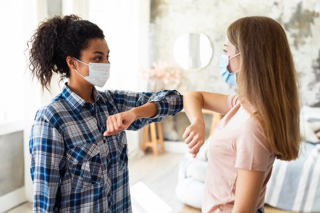 Seitenansicht von Freundinnen mit medizinischen Masken, die den Ellbogengruß üben