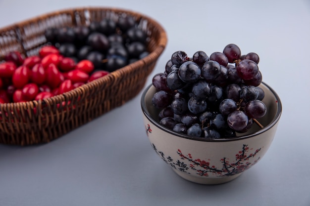 Seitenansicht von frischen dunkelhäutigen Trauben auf einem Eimer mit Kornelkirschenbeeren auf einem grauen Hintergrund