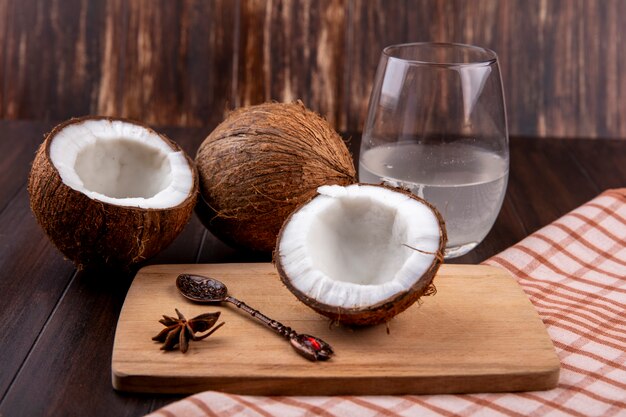 Seitenansicht von frischen Kokosnüssen auf einem hölzernen Küchenbrett mit Löffel und einem Glas Wasser auf karierter Tischdecke und Holzoberfläche