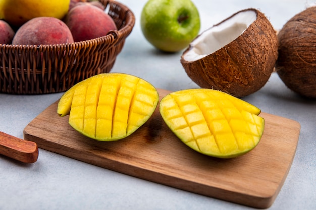 Seitenansicht von frischen und köstlichen Früchten wie geschnittener Mango auf einem hölzernen Küchenbrett mit Pfirsichen auf einem Eimer grüner Apfel und halber Kokosnuss auf weißer Oberfläche