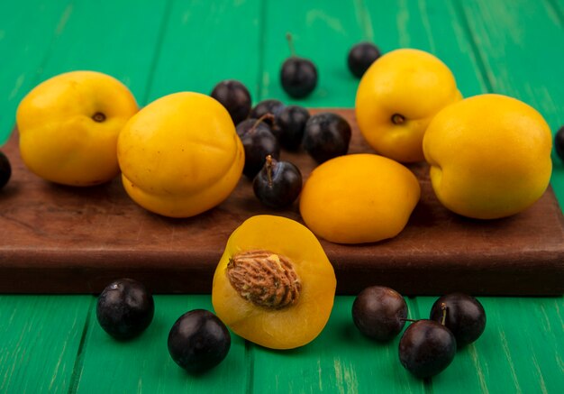 Seitenansicht von Früchten als Aprikosen und Schlehenbeeren auf Schneidebrett mit halber Aprikose auf grünem Hintergrund