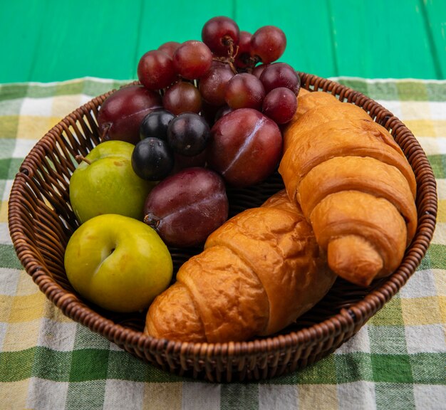 Seitenansicht von Früchten als Traubenpluots-Schlehenbeeren mit Croissants im Korb auf kariertem Stoff und grünem Hintergrund