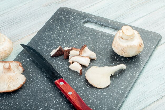 Seitenansicht von ganzen und geschnittenen frischen Pilzen mit Küchenmesser auf einem schwarzen Schneidebrett auf rustikalem Tisch