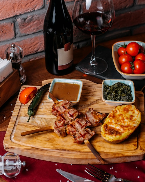 Seitenansicht von gegrilltem Hühnerfleisch auf Holzstäbchen mit Ofenkartoffel und Soße auf einem Holzbrett