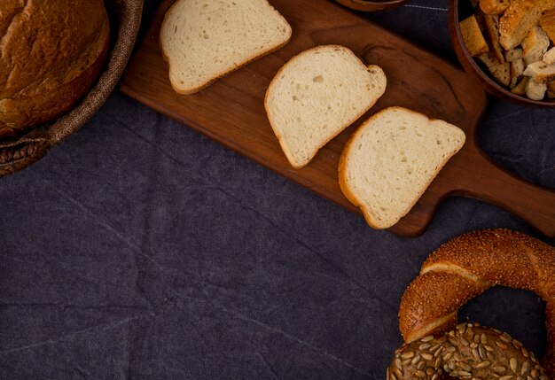 Seitenansicht von geschnittenem Weißbrot auf Schneidebrett mit Bagels und Brotstücken auf kastanienbraunem Hintergrund mit Kopienraum