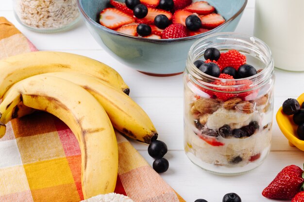 Seitenansicht von Hafer über Nacht mit frischen Erdbeeren, Blaubeeren und Nüssen in einem Glas und reifen Bananen auf rustikalem Tisch