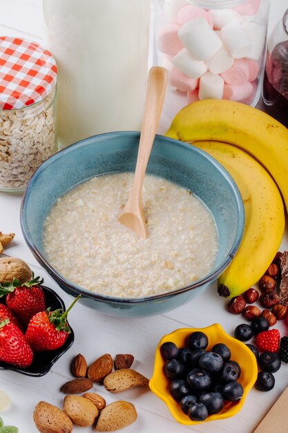 Seitenansicht von Haferbrei in einer Schüssel und frischen Beerenbananen und -nüssen auf weißem rustikalem Tisch