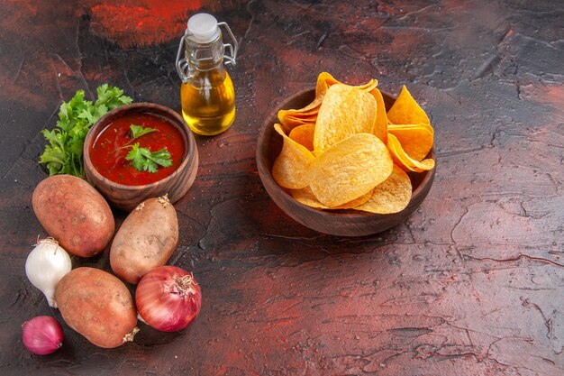 Seitenansicht von hausgemachten leckeren knusprigen Chips in einer kleinen braunen Schüssel Knoblauch grüne Ketchup-Kartoffel-Zwiebel-Öl-Flasche auf dunklem Tisch