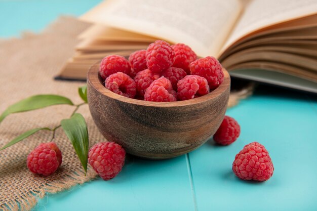 Seitenansicht von Himbeeren in Schüssel und offenem Buch mit Blättern auf Sackleinen und blau