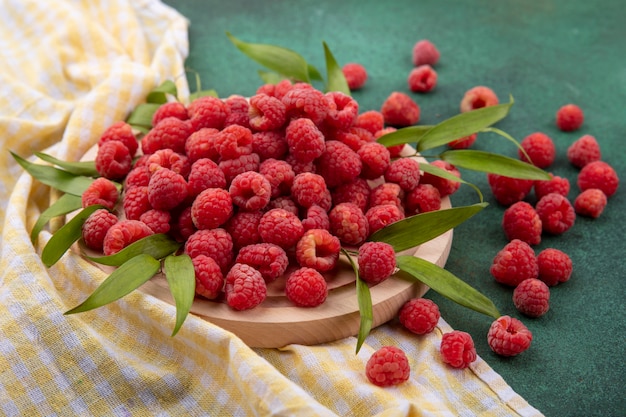 Kostenloses Foto seitenansicht von himbeeren mit blättern auf schneidebrett auf kariertem stoff und auf grün