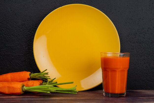 Seitenansicht von Karottensaft und Karotten mit leerem Teller auf Holzoberfläche und schwarzem Hintergrund