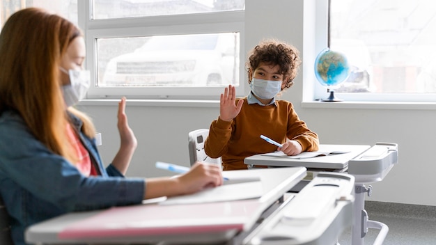 Seitenansicht von Kindern mit medizinischen Masken im Klassenzimmer, die sich aus der Ferne grüßen