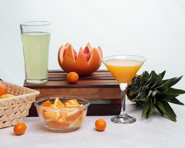 Seitenansicht von Limettensaft im Glas und geschnittener Grapefruit mit Kumquat auf Schneidebrett mit Orangenscheiben in Schüssel Ananas und Glas Orangensaft auf weißem Hintergrund