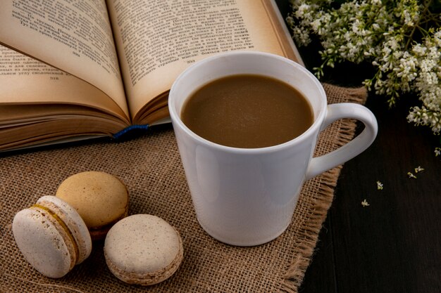 Seitenansicht von Macarons mit einer Tasse Kaffee auf einer beigen Serviette mit einem offenen Buch und Blumen auf einer schwarzen Oberfläche