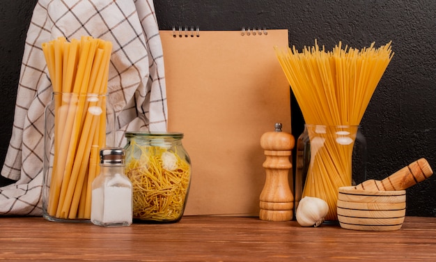 Seitenansicht von Makkaroni als Bucatini und Spaghetti in Gläsern mit Salz-Knoblauch-Knoblauch-Brechertuch und Notizblock auf Holzoberfläche und schwarzem Hintergrund mit Kopienraum