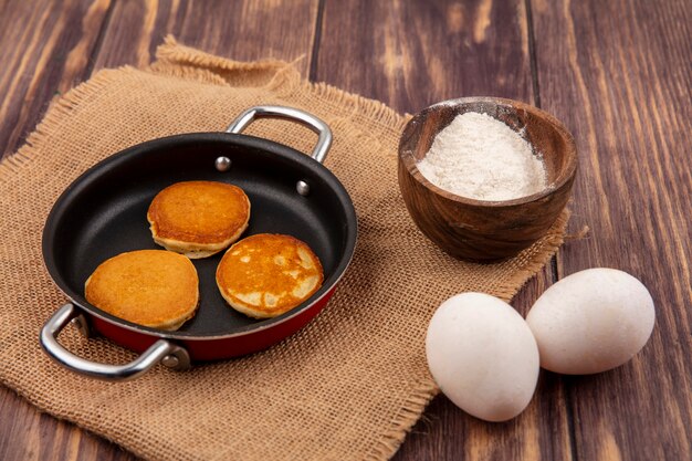 Seitenansicht von Pfannkuchen in Pfanne und Schüssel Mehl auf Sackleinen mit Eiern auf hölzernem Hintergrund