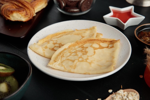 Seitenansicht von Pfannkuchen in Teller mit Croissant-Kiwi-Plätzchen-Marmelade-Hafer auf schwarzem Hintergrund