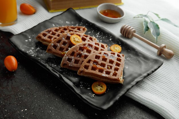 Seitenansicht von Pfannkuchen mit Kumquat-Scheiben in Teller und Marmelade-Orangensaftblättern auf Stoff auf schwarzem Hintergrund