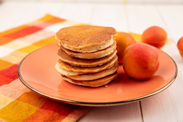 Seitenansicht von Pfannkuchen und Aprikosen im Teller auf kariertem Stoff und hölzernem Hintergrund