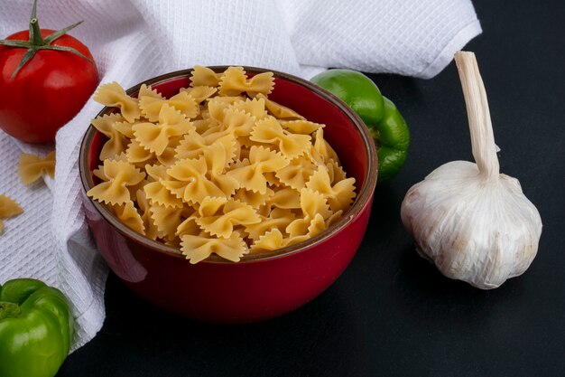 Seitenansicht von rohen Nudeln in einer Schüssel mit Tomaten Knoblauch und Paprika auf einem weißen Handtuch auf einer schwarzen Oberfläche