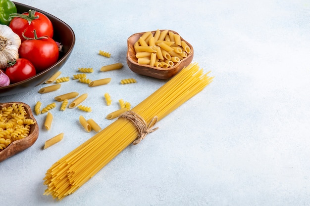 Seitenansicht von rohen Spaghetti mit rohen Nudeln in einer Schüssel mit Tomaten und Knoblauch in einer Schüssel auf einer grauen Oberfläche