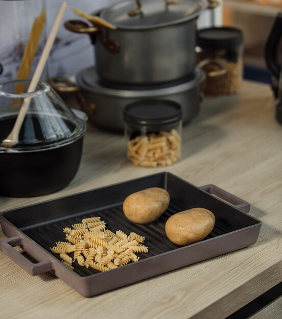 Seitenansicht von rohen Spiralnudeln und zwei Kartoffeln in einer Pfanne auf einem Küchentisch