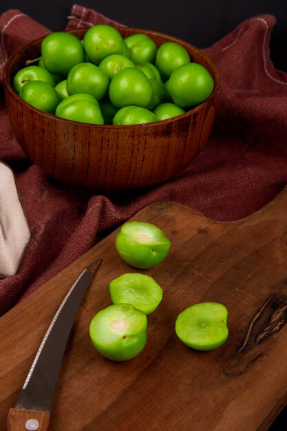 Seitenansicht von sauren grünen Pflaumen in einer Holzschale und geschnittenen grünen Pflaumen mit einem Küchenmesser auf einem Holzbrett auf dunkelrotem Stofftisch
