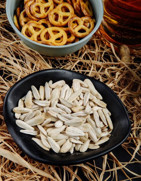 Seitenansicht von Sonnenblumenkernen in einer schwarzen Schüssel mit Mini-Brezeln und einem Becher Bier auf Stroh