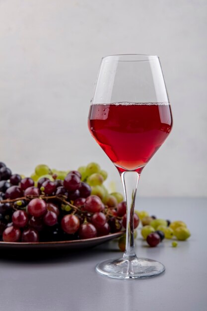 Seitenansicht von Traubensaft in Weinglas und Trauben in Platte und auf grauer Oberfläche und weißem Hintergrund