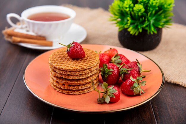 Seitenansicht von Waffelkeksen und Erdbeeren in Teller und Tasse Tee mit Zimt auf Untertasse und Blume auf Sackleinen und Holz